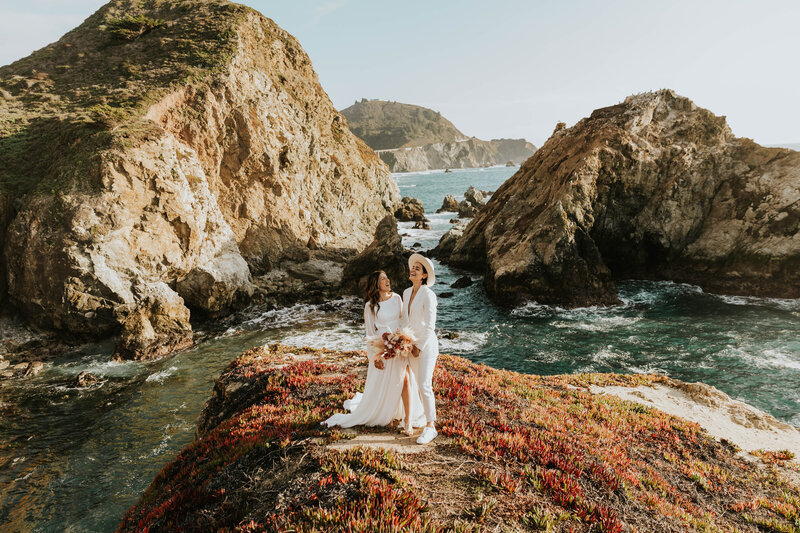 two brides standing on cliff next to ocean laughing