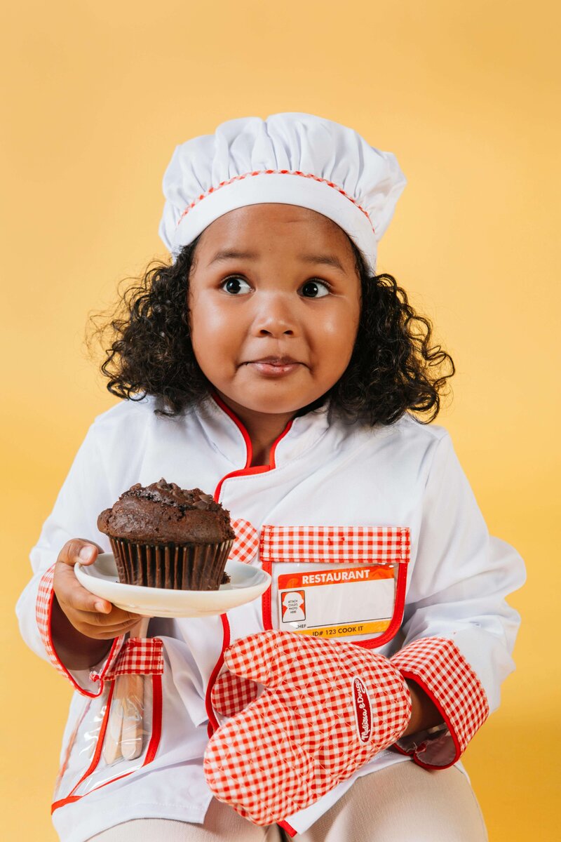 A young girl dressed as a chef, wearing a white hat and red-checkered apron, holds a chocolate muffin on a plate, standing against a bright yellow background.