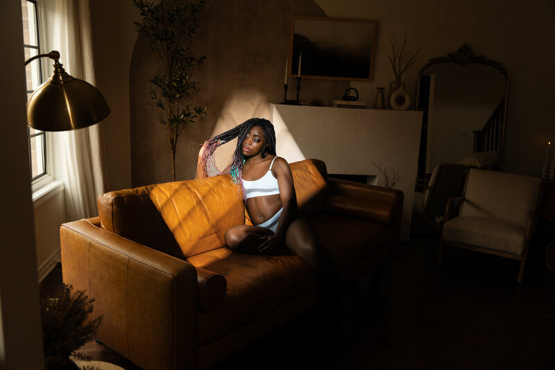 black woman with braids in a white sports bra and undies sitting on a couch with harsh window light marks lighting her