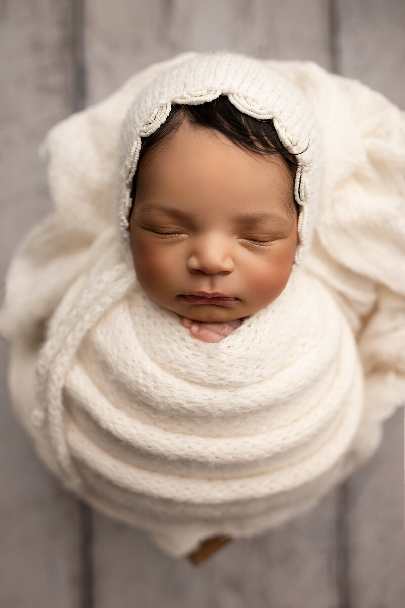 Baby is swaddled in cream with a matching bonnet with scalloped detail for a NJ newborn photoshoot. Aerial image. Baby is sleeping with her hands peeking out from the swaddle and resting under her chin. Captured by top NJ newborn photographer Katie Marshall.