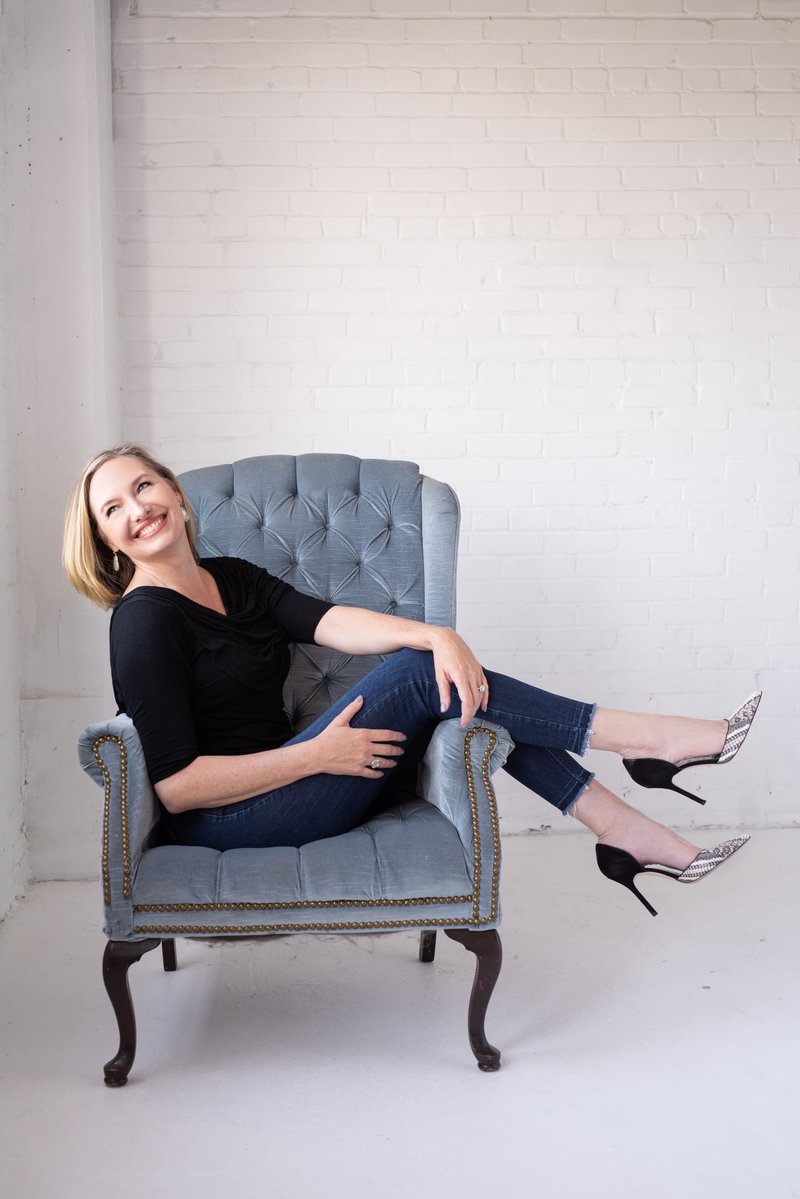Headshot of Jen Denton sitting with high heels and smiling looking out a window