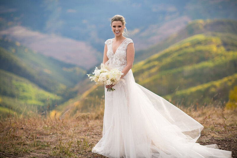 Testimonial photo, bride in flowing lace dress smiles at a camera with hills behind her