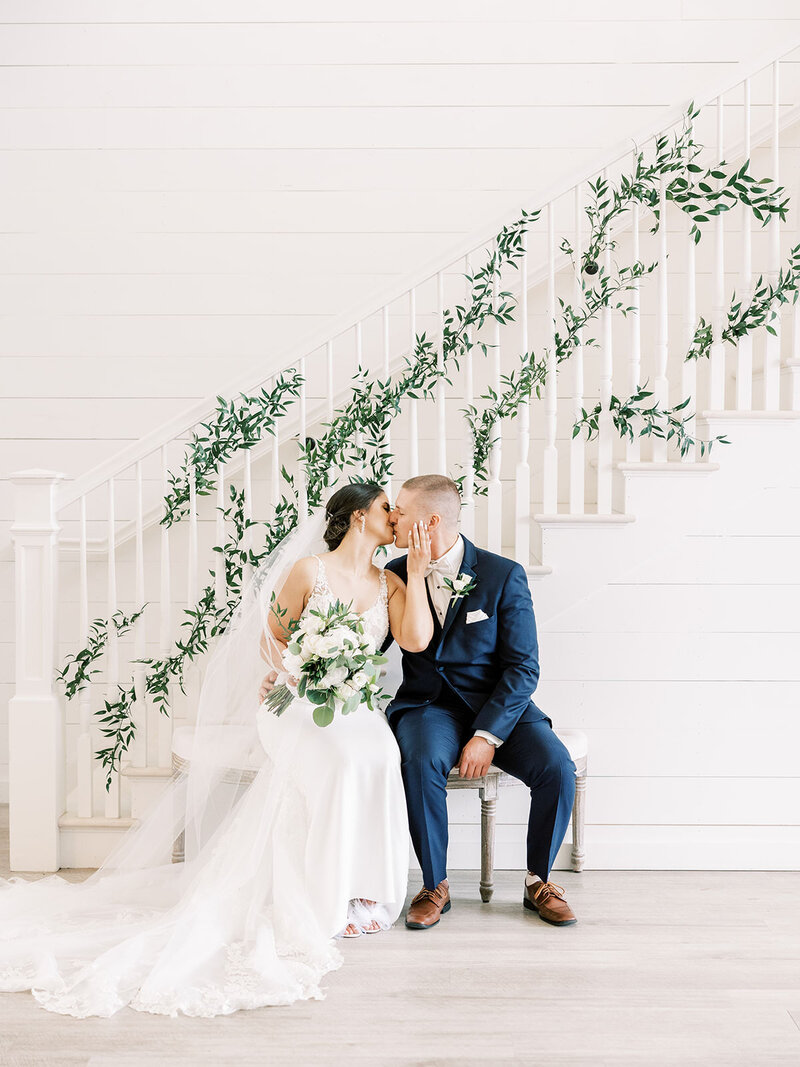 Bride and groom kiss at the base of a white staircase covered in vines on their wedding day