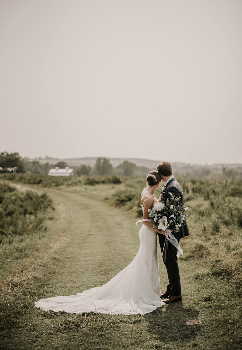 An image of a bride and groom on their wedding day, standing in a field looking off into the horizon.