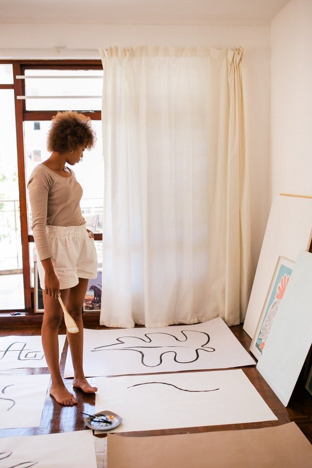 women standing with bare feet and a big paintbrush in hand looking at her own paintings on the floor