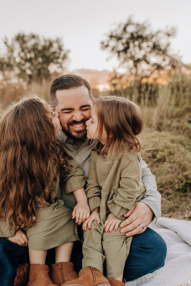 A family mini session photoshoot in a grassy San Antonio field. Two toddler girls are kissing dad on each cheek while he smiles.