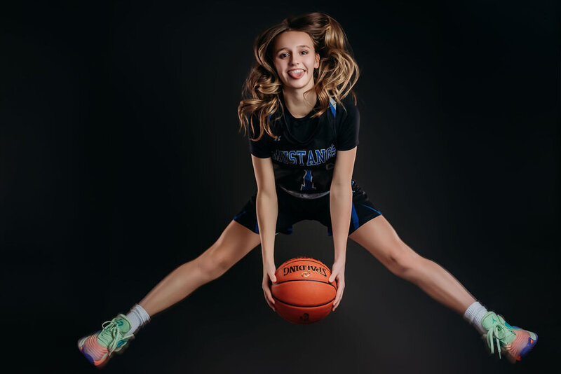 A teenage girl jumps with a basketball in front of a blue solid backdrop in an action shot for her senior graduation photoshoot, captured by a fine art studio photographer in Bemidji, Minnesota