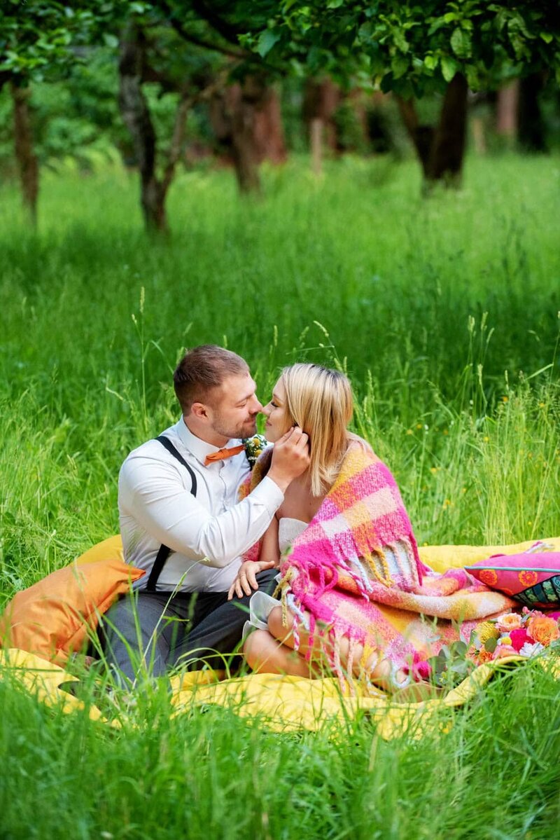 bride wrapped in a colorful blanket touches noses with a groom while sitting in bright green tall grass