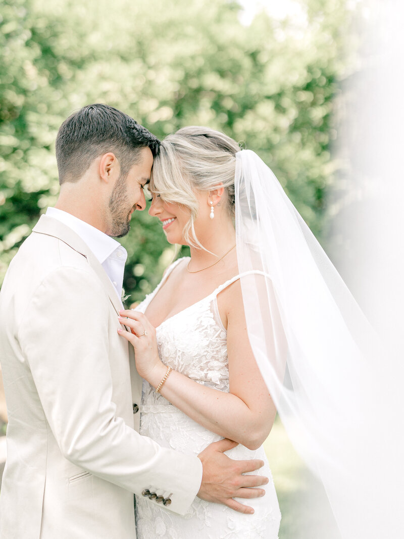 Bride wearing veil and groom in a tan suit standing with their foreheads together