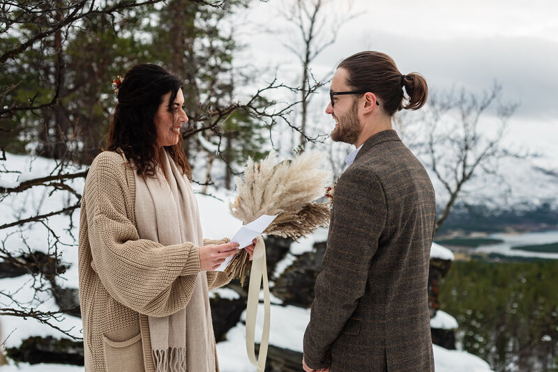 A woman and a man are standing facing each other against a snowy winter landscape, during their elopement ceremony. The woman, wearing a cream shawl and a dress, is holding a paper in one hand and a bouquet made of pampas grass and flowers in the other, reading her vows. Adorned with delicate flowers in her hair, she looks at the man conveying her love for him in every sense. The man, with his hair tied back and wearing a herringbone-patterned tweed suit, listens attentively. Behind them, bare trees and a glimpse of a frozen lake set a serene and natural backdrop, for their wedding ceremony.