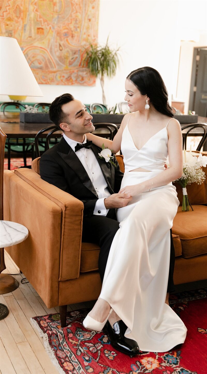 Woman with long, black hair and simple wedding gown sits on groom's lap