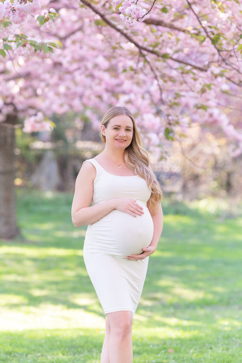 A pregnant women wearing a beige colored dress holding her belly at a park with beautiful pink flower bloom in the background.
