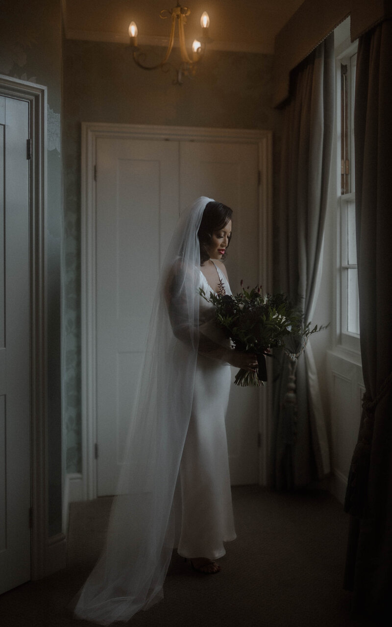Bride standing by the window with her bouquet, bathed in natural light, capturing a moment of grace and beauty as she prepares for her wedding day