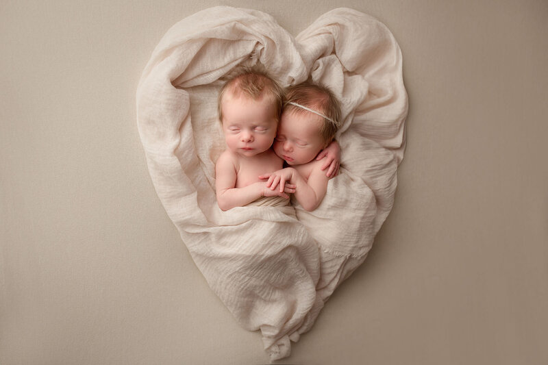 twin sisters posed on a cream blanket with a wrap shaped as a heart holding each other