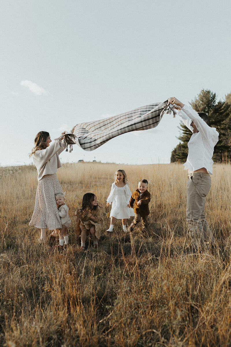 Capturing the essence of a family photoshoot in a picturesque field, where parents spread a blanket and children joyfully play beneath it.