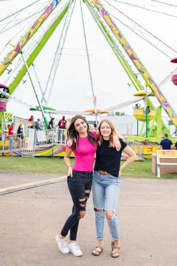 Two young women smiling and standing in front of a ferris wheel at a fairground.