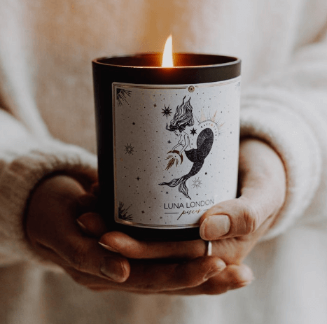 woman holding a lit candle in the palm of her hands, the candle has an illustration on the front of the zodiac sign Pisces