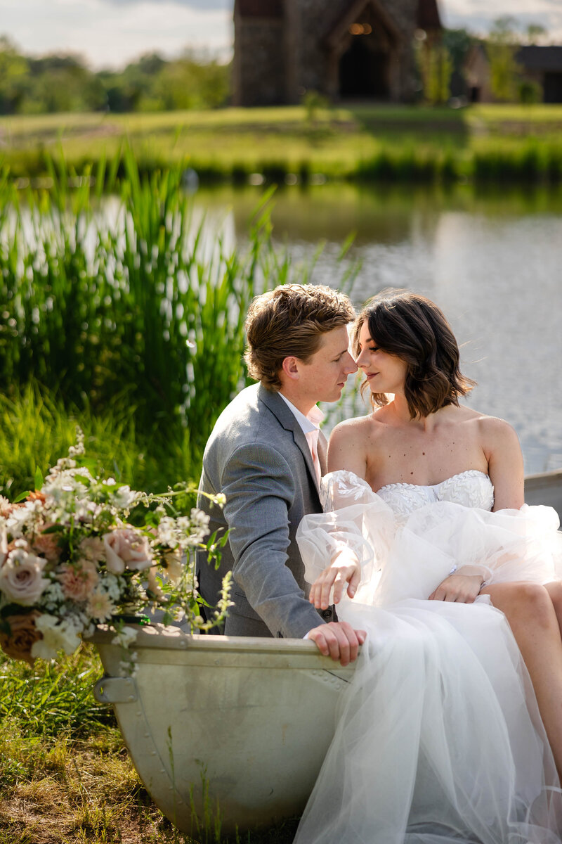 Bride and Groom sitting in a row boat on a pond with florals cascading down front of the boat.  Bride is dressed in a strapless fitted bodice gown with separate lace sleeves and full skirt  Groom is in a gray suit.  Bride and groom are looking at each other while sitting in the boat.