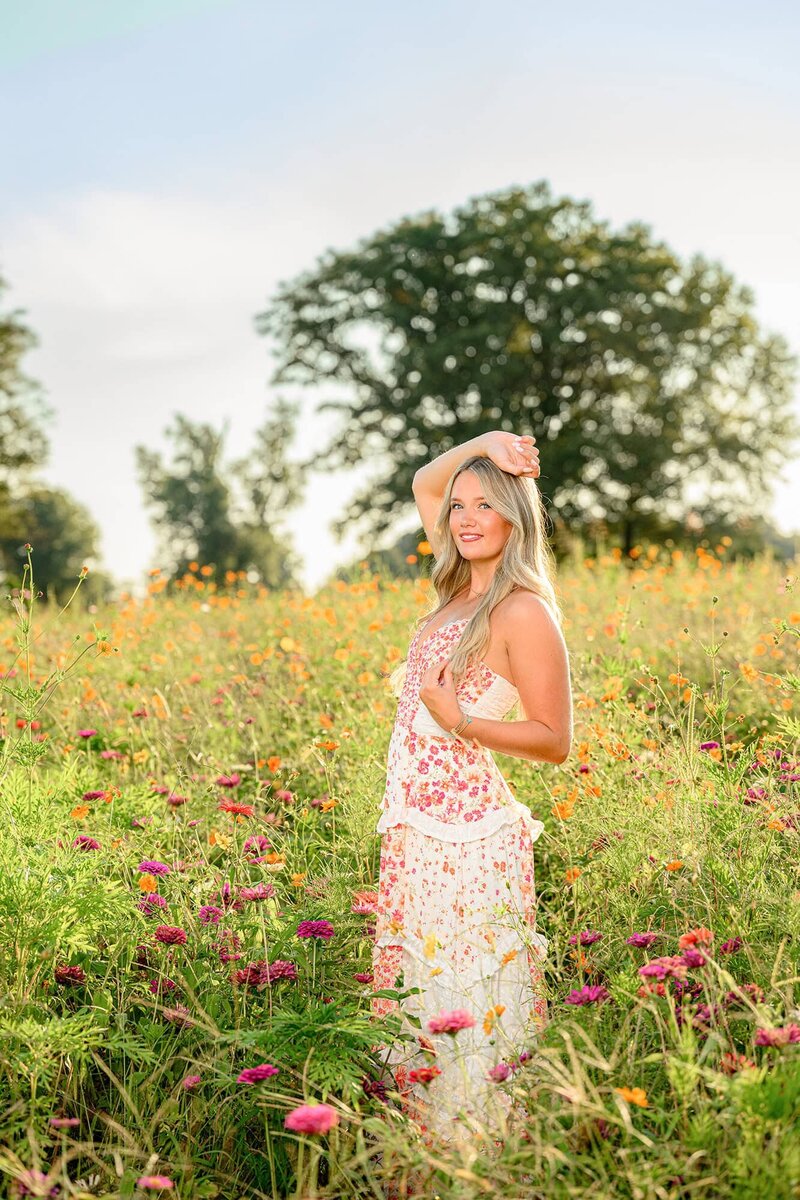 A senior girl with long blonde hair stands in a floral field wearing a pink and orange floral dress