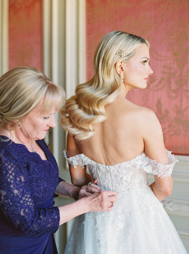 A mother in a blue lace dress helping her daughter, the bride, put on her strapless wedding gown at The Olana in Dallas Texas