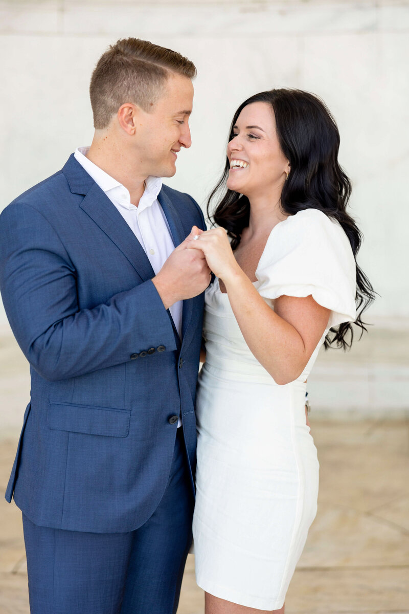 A couple smiling and holding hands. The man is wearing a blue suit with a white shirt, and the woman is in a white dress. They are indoors against a light-colored background.