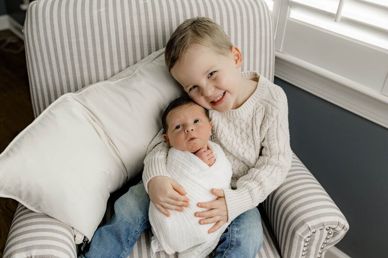 Brother holds his newborn brother wrapped and cream while he's dressed in a cream sweater and jeans sitting in a chair