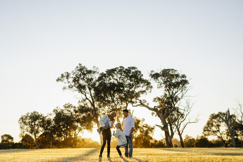lifestyle-family-photographer-perth-002 Wide-angle silhouette portrait of a family standing in an open field surrounded by rustic trees during a warm sunrise photography session in Perth.