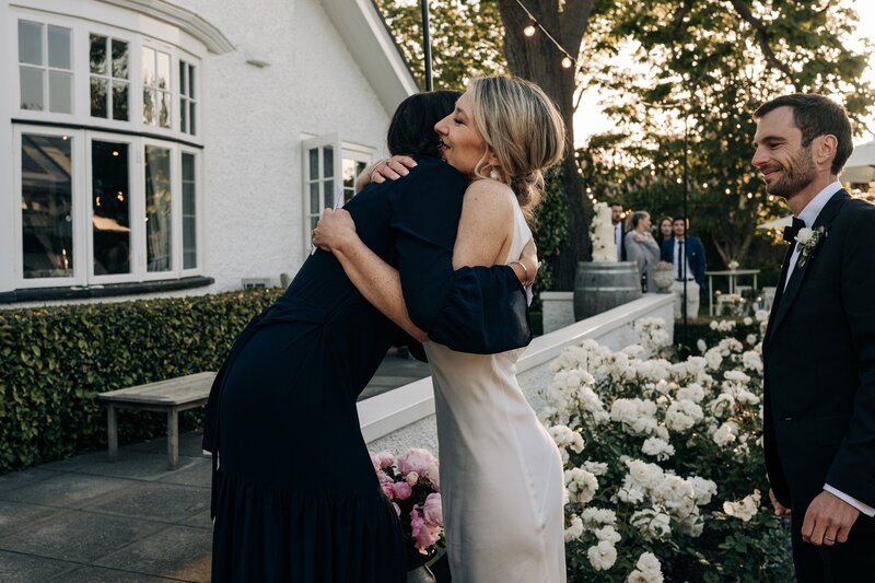 a bride hugs a friend in candid moment with white roses behind and sunset
