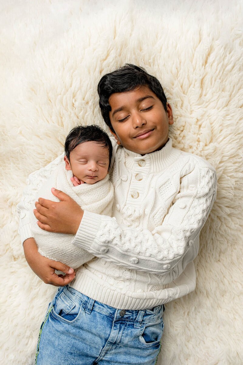 Older brother is dressed in a cream sweater with his newborn baby brother wrapped in cream laying beside him on a fur rug both of their eyes are closed