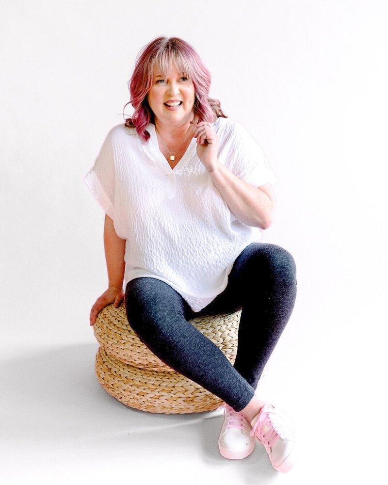 woman with pink hair sits on stool and laughs, wearing white shirt and black leggings