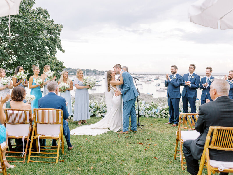 Landscape photo of bride and groom kissing during wedding ceremony while bridal party cheers