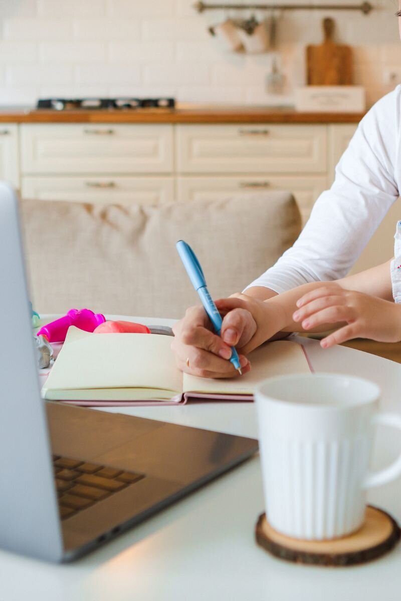 Mom entrepreneur writing in a notebook with a child’s hand nearby and a laptop on the table.