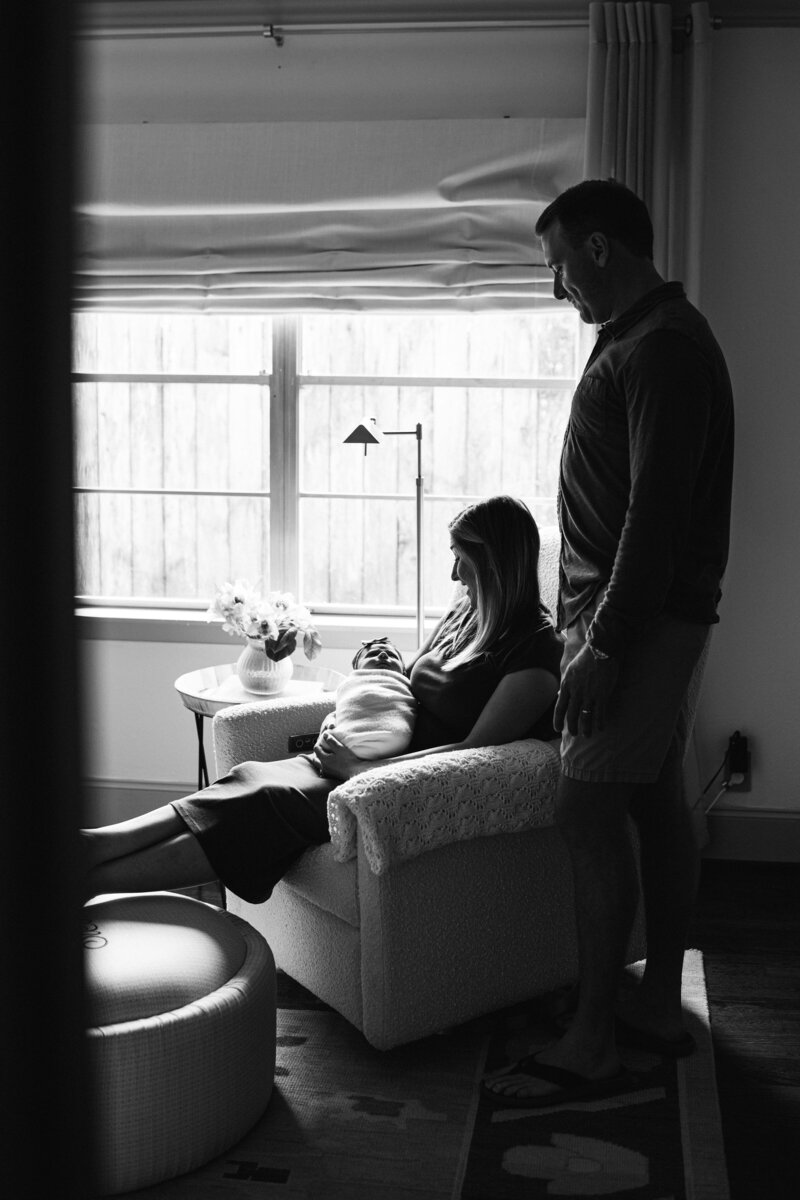 Mother sitting holding newborn, father standing behind and gazing lovingly