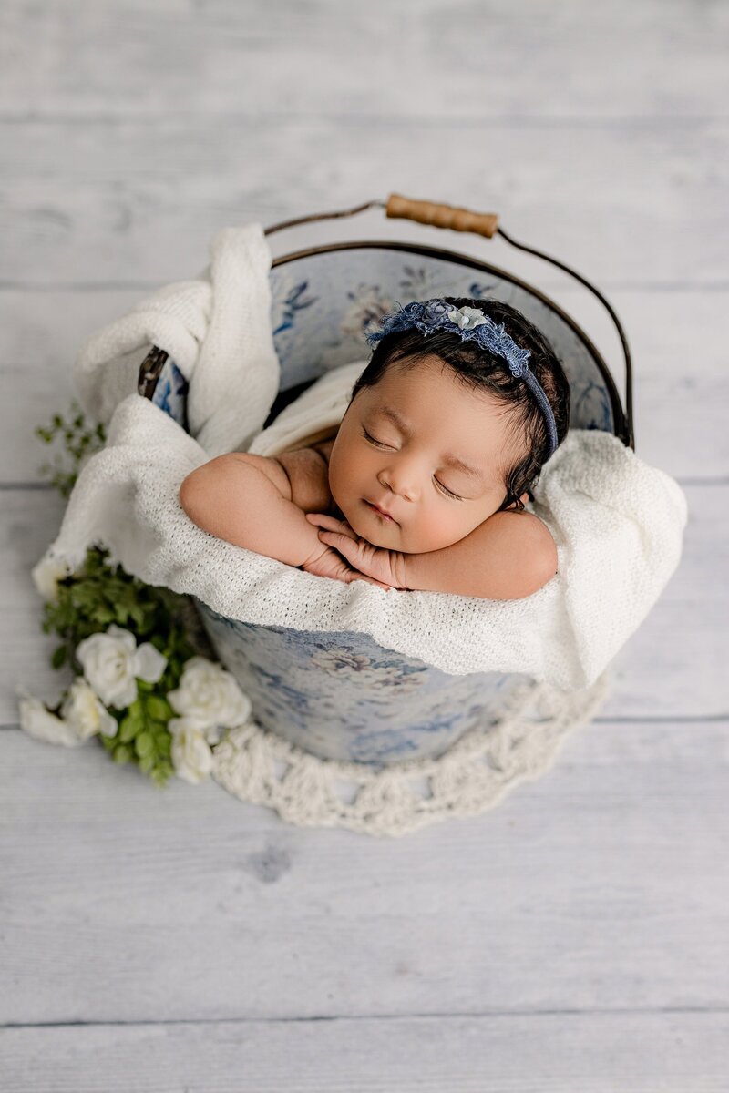 Newborn baby girl is placed in a bucket with blue flowers laying on a cream layer white knit layer and wearing a blue flower headband in a Charlotte Area Studio
