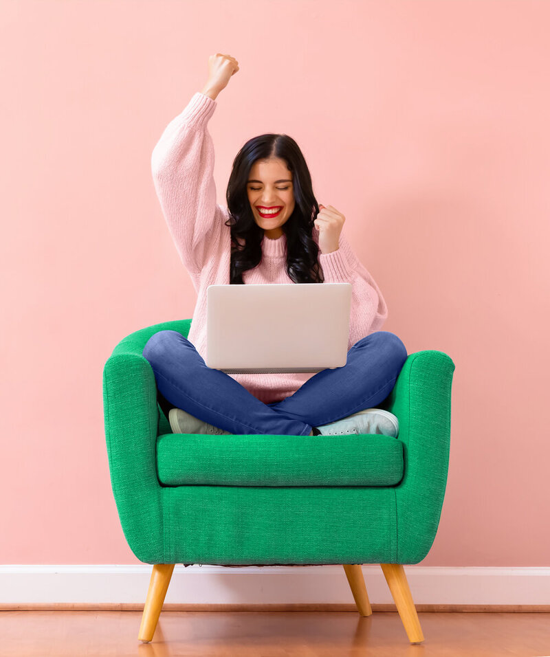 Young female entrepreneur sitting on a green  sofa chair with crossed legs and a laptop and  raising her hands  in the air.
