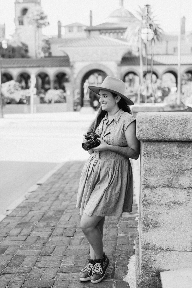 A candid black and white photo of Orlando Wedding Photographer Jo from Four Loves Photo and Film. She is wearing a short sleeved shirt dress and wide brimmed hat, leaning casually against a brick pillar on a cobblestone sidewalk.