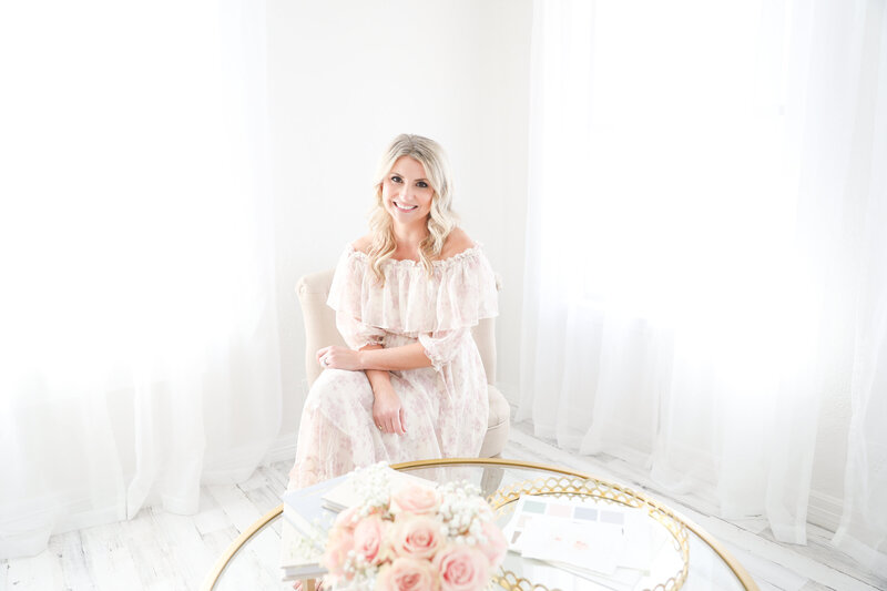 Austin Newborn Photographer Rebecca Dyan headshot sitting in white studio on cream chair wearing a floral dress smiling at the camera