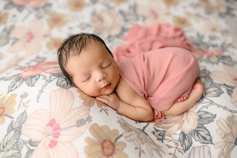 newborn baby girl is covered in a pink wrap while posed on a flower background