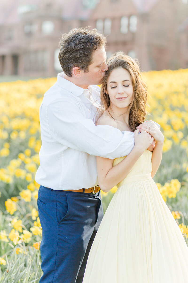 Man and woman are standing in a field of daffodils for their Ipswitch engagement photoshoot . Woman is in a pale yellow gown with her back to her fiance. The man has his arms wrapped around the woman's chest and he is kissing her atop of her head. Captured by top Ipswitch wedding photographer Lia Rose Weddings