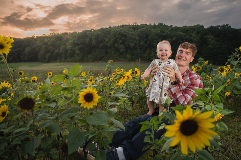 A dad holding a baby smiling in a patch of sunflowers near Boston