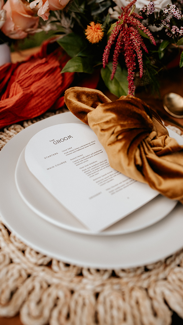 White curved menu card on a plate with a brown velvet napkin and flowers