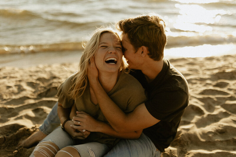 A couple sitting in the sand and holding each other while laughing