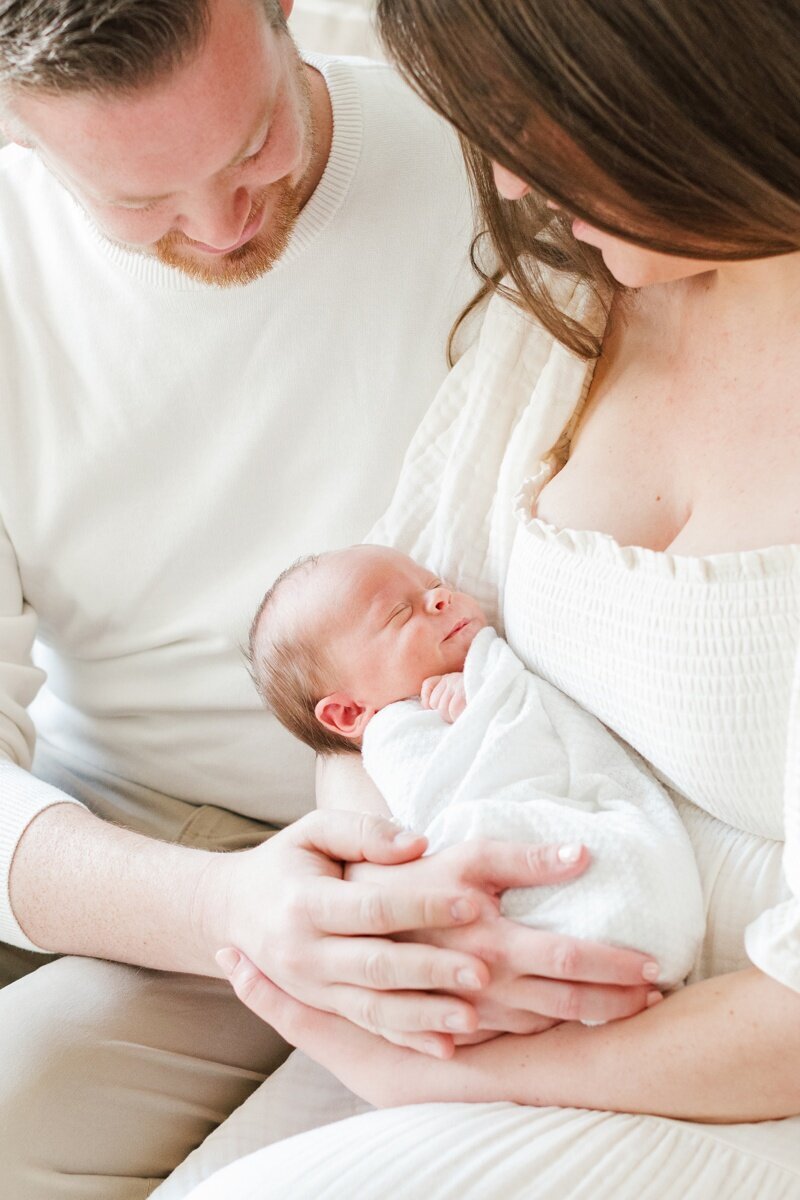 A couple snuggles their newborn baby while being photographed by New Jersey Portrait Photographer Kate Voda. The husband is wearing a white sweater and khaki pants, the wife is wearing a cream color dress. The baby is wrapped in a white swaddle.