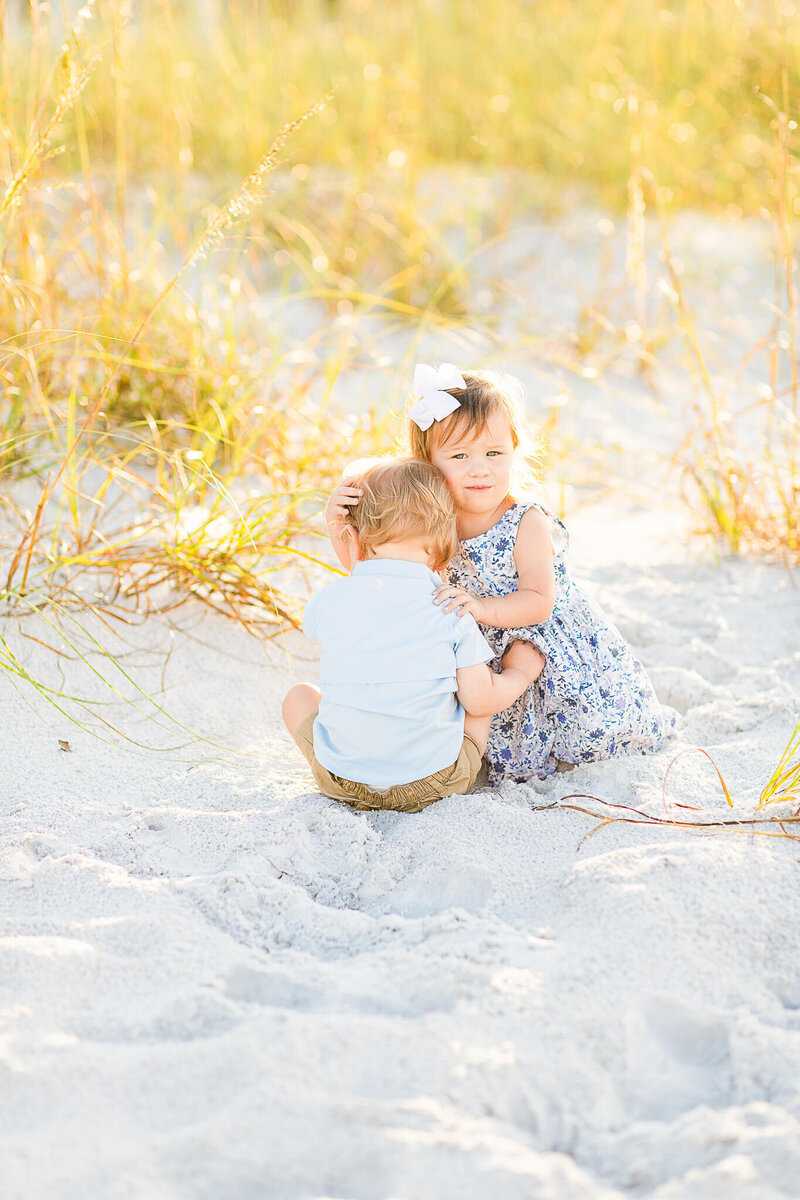 Little girl in a floral dress hugs little boy in a blue shirt at the beach