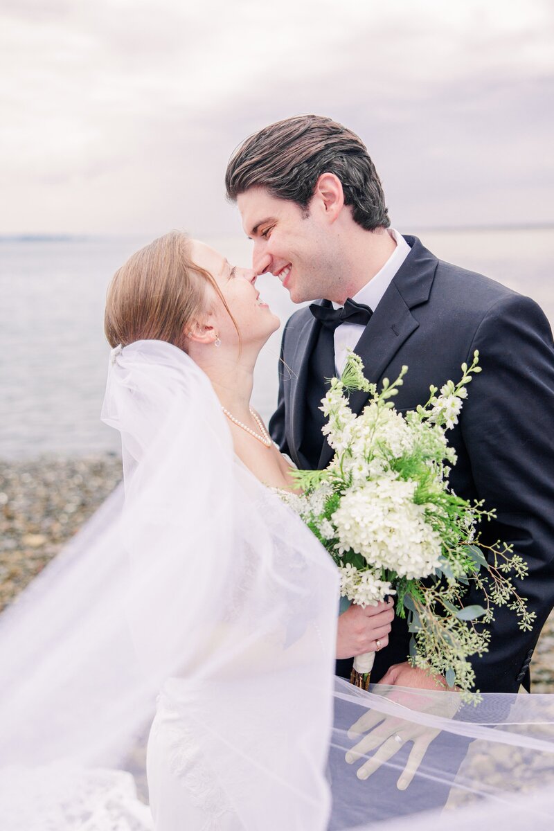 Bride and groom touching noses on the beach with veil blowing in the breeze