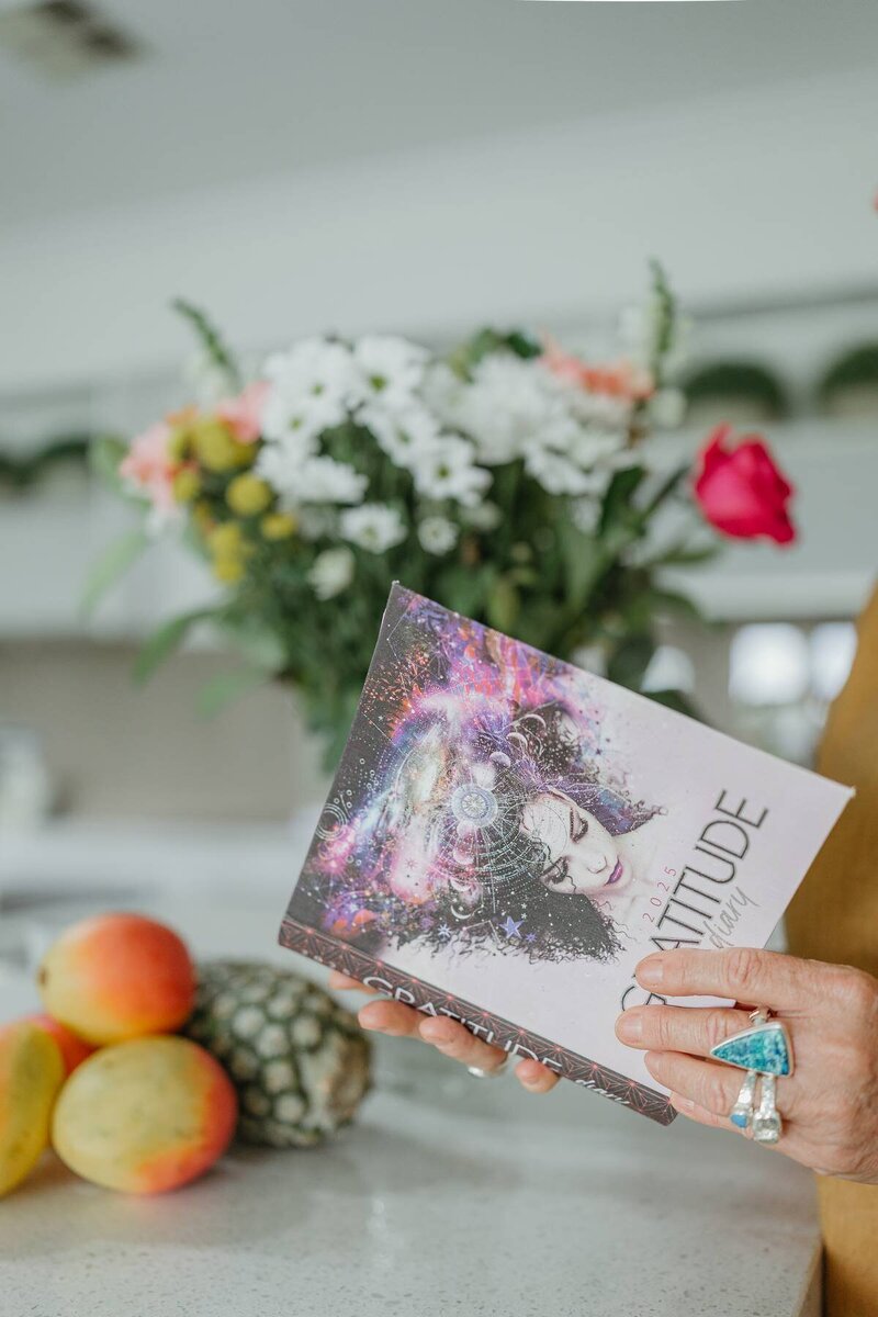 Branding photography capturing a business owner holding a colourful gratitude journal beside fresh produce and flowers.