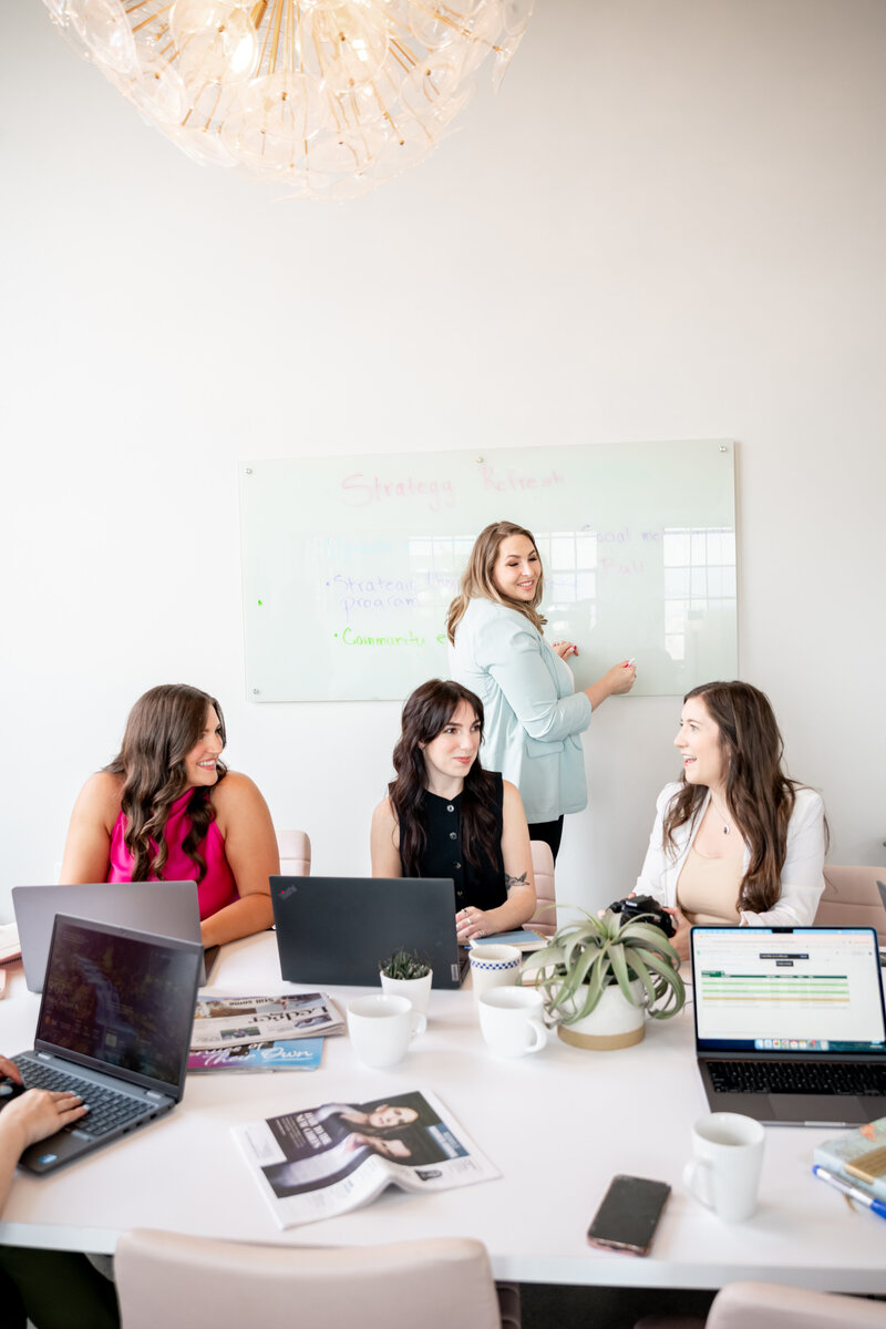Four Pierce PR team members collaborate at a table, smiling and discussing a project on a laptop.