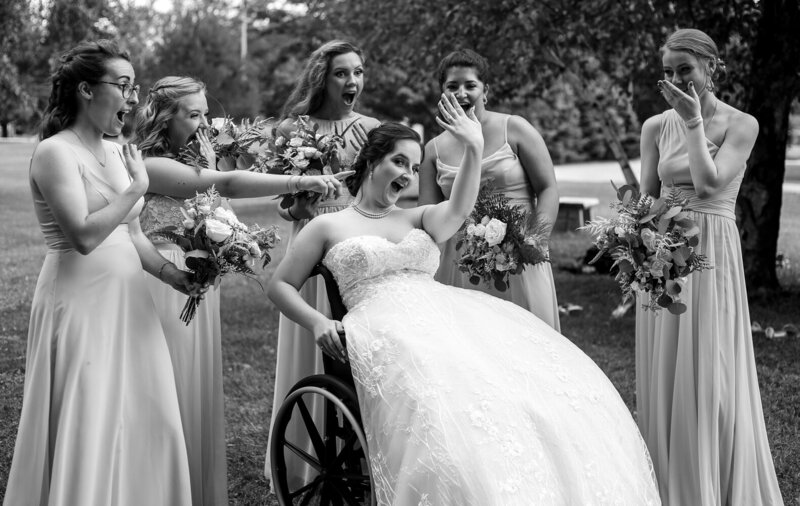 Bride sits in wheelchair surrounded by bridesmaids and shows off her new wedding rings while they happily react in awe