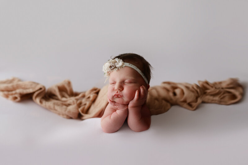 newborn baby girl with head resting on hands in froggy pose sleeping with white background and pretty bow