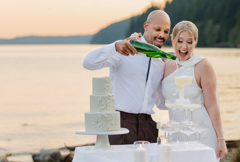 Groom pours the champagne bottle on to the champagne tower. Bride is happy and excited to see champagne fall. Lake and sun set in the back round.